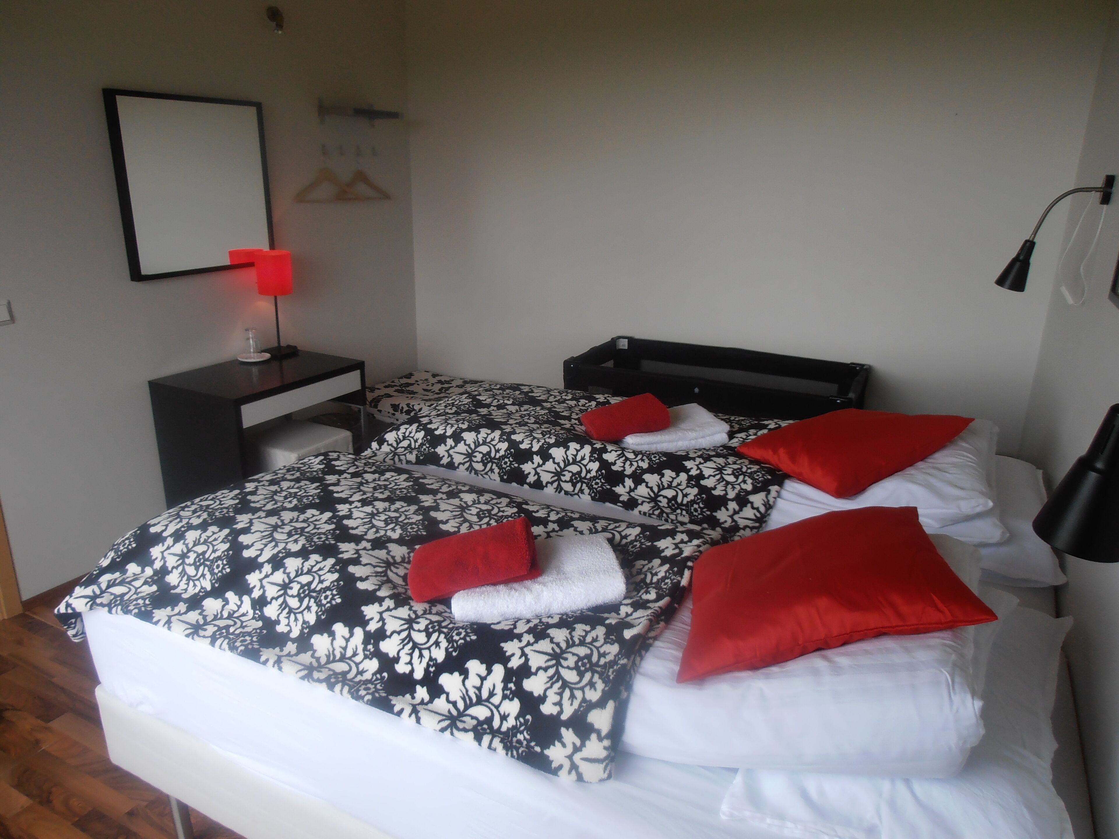Een hotelkamer met twee bedden, elk met een zwart-wit bloemenbedekking, rode kussens en witte handdoeken. Er is een bureau met een rode lamp, een zwarte stoel en een spiegel aan de muur.
