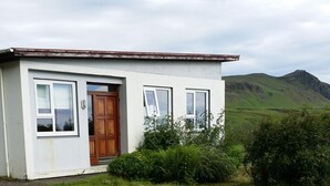 A single-story house with a white exterior, a wooden door, and a small front yard with grass and bushes. - Giljur Guesthouse (Vik I Myrdal)