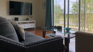 Een moderne woonkamer met een bank, een glazen salontafel, een flatscreen-televisie aan de muur en uitzicht op een balkon met groen.