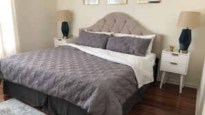 Een netjes opgemaakt bed met een grijze deken, twee nachtkastjes met lampen en een ingelijst schilderij aan de muur.