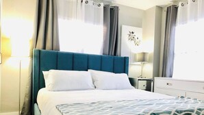 Un lit bien fait, avec du linge de lit blanc et une tête de lit bleue, une table de chevet avec une lampe, et une vue sur l’extérieur à travers des rideaux légers.