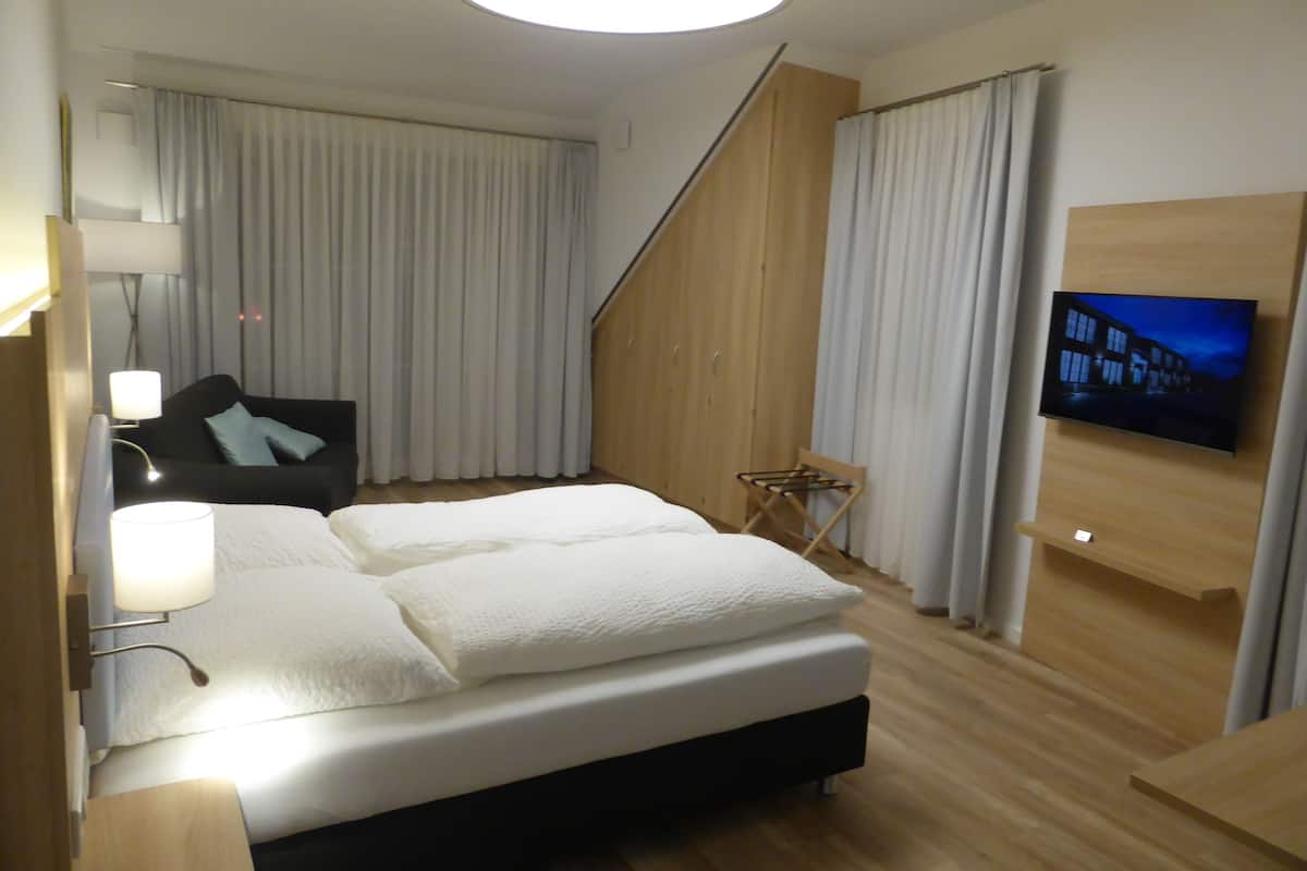 A modern hotel room with a large bed, a flat-screen TV mounted on the wall, a wooden wardrobe, and a small seating area with a chair and table.