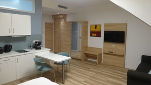 A modern kitchen with white cabinets, a coffee machine, and a water bottle. There is a dining area with a white table and blue chairs. A black sofa and a TV are also visible. - Leister Apparthotel (Weyhe)