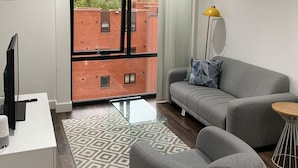 Ein modernes Wohnzimmer mit einem grauen Sofa, einem kleinen runden Tisch, einem Fernseher auf einem weißen Schrank und einem gemusterten Teppich.
