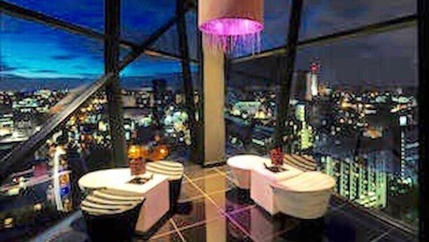 Ein Hotelzimmer in einem Hochhaus mit Panoramablick auf die Stadt, zwei runden Tischen und einer großen Hängelampe.