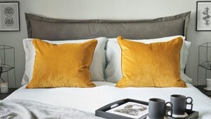 Ein ordentlich bezogenes Bett mit zwei senffarbenen Kissen, einem Tablett mit zwei Tassen und einem gerahmten Bild daneben.