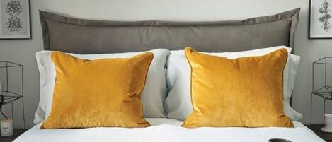 Een net opgemaakt bed met twee mosterdgele kussens, een dienblad met twee mokken en een ingelijste foto aan de zijkant.