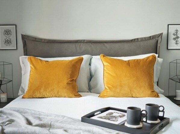 A neatly made bed with two mustard pillows, a tray with two mugs, and a framed picture on the side.