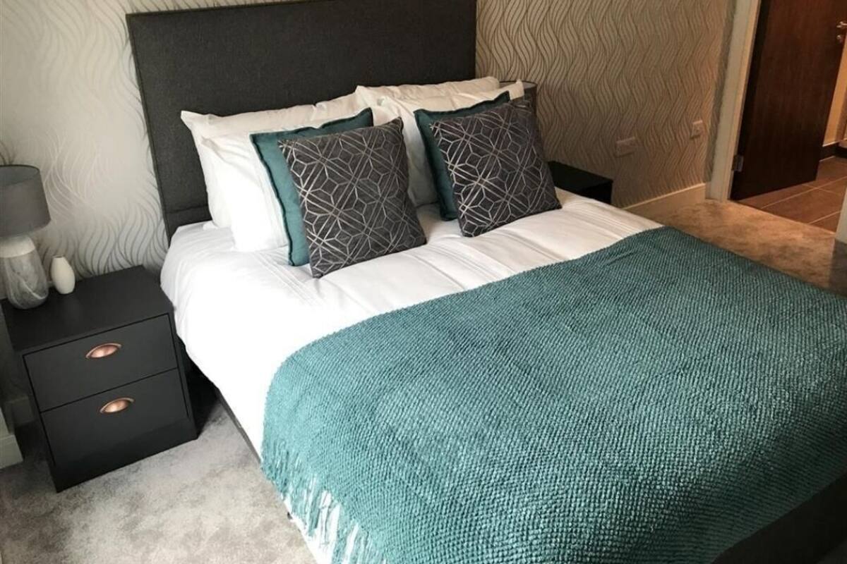 A neatly made bed with a white comforter and teal throw blanket, a dark headboard, and a nightstand with drawers.