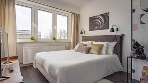 A bedroom with a large bed, a window with curtains, a radiator, a small plant, and a statue on a shelf. - Studio des roses 4 * place des Martyrs hyper center (Luxembourg)