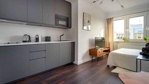 Una cocina moderna con armarios grises, horno integrado y cafetera. Hay una mesita de madera con un televisor y una cama con edredón blanco.