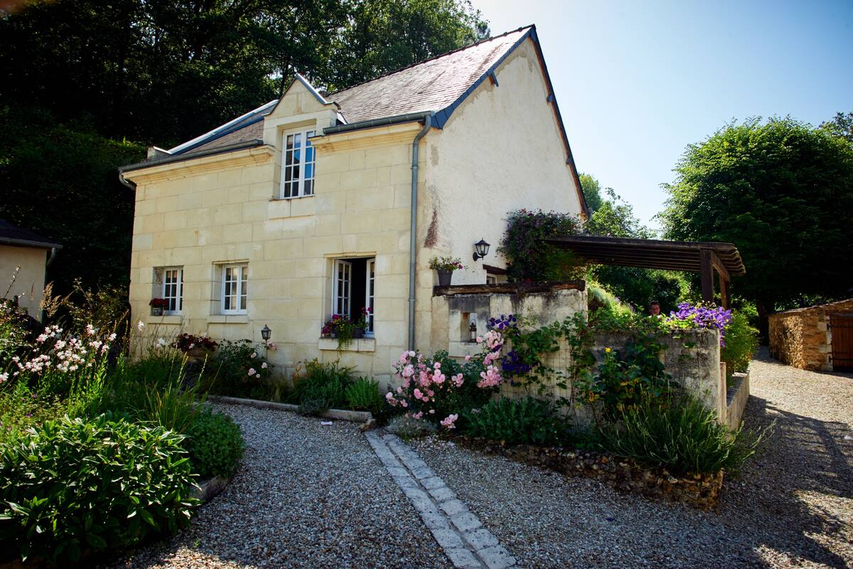 A two-story stone house with a gravel driveway, surrounded by lush greenery and flowering plants.