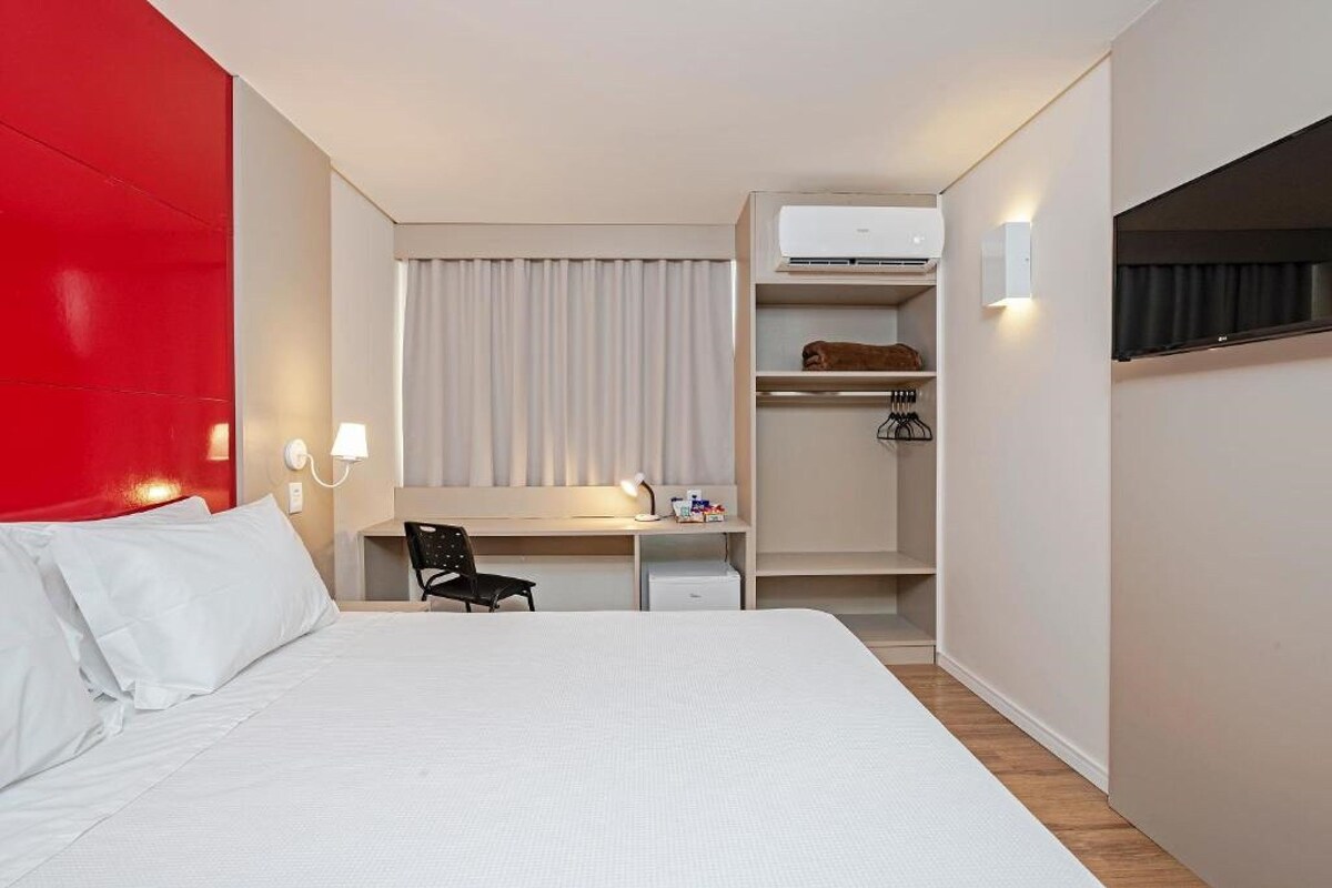 A hotel room with a bed, a desk, a chair, a wardrobe, and an air conditioning unit.