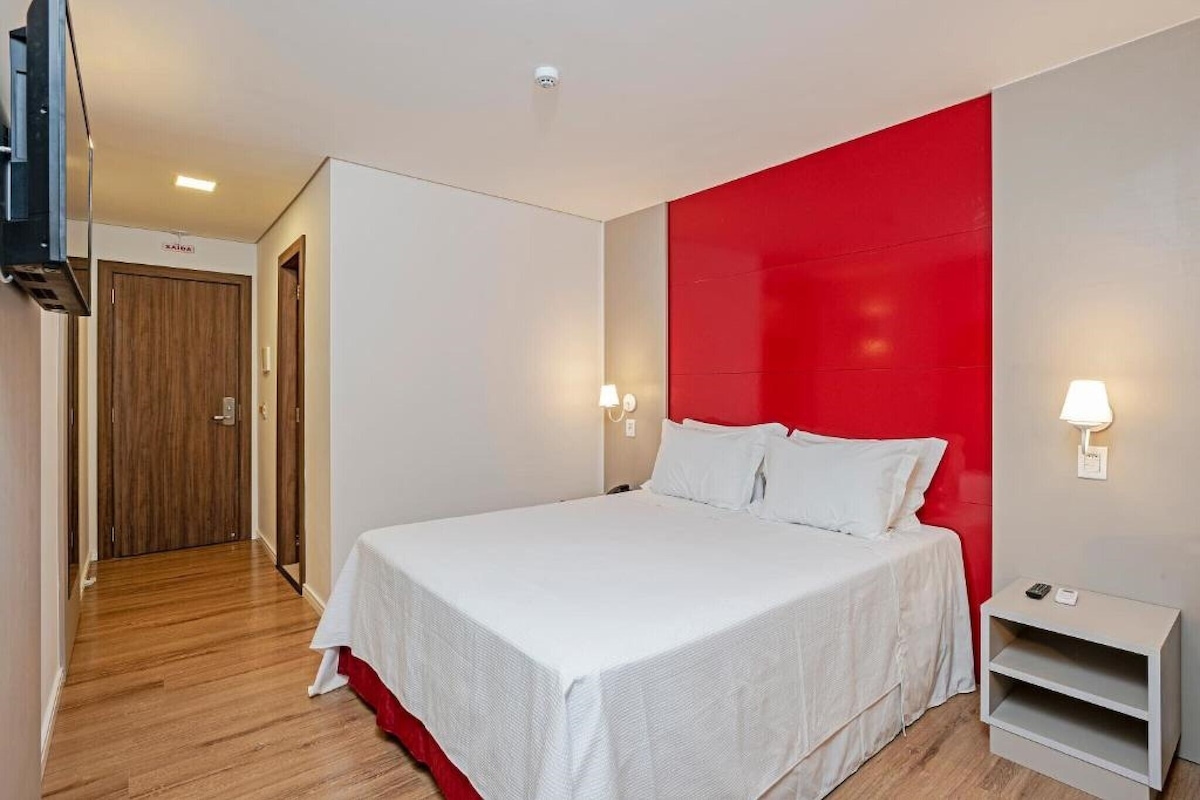 A modern hotel room with a large bed, a red accent wall, and a small bedside table with a lamp.