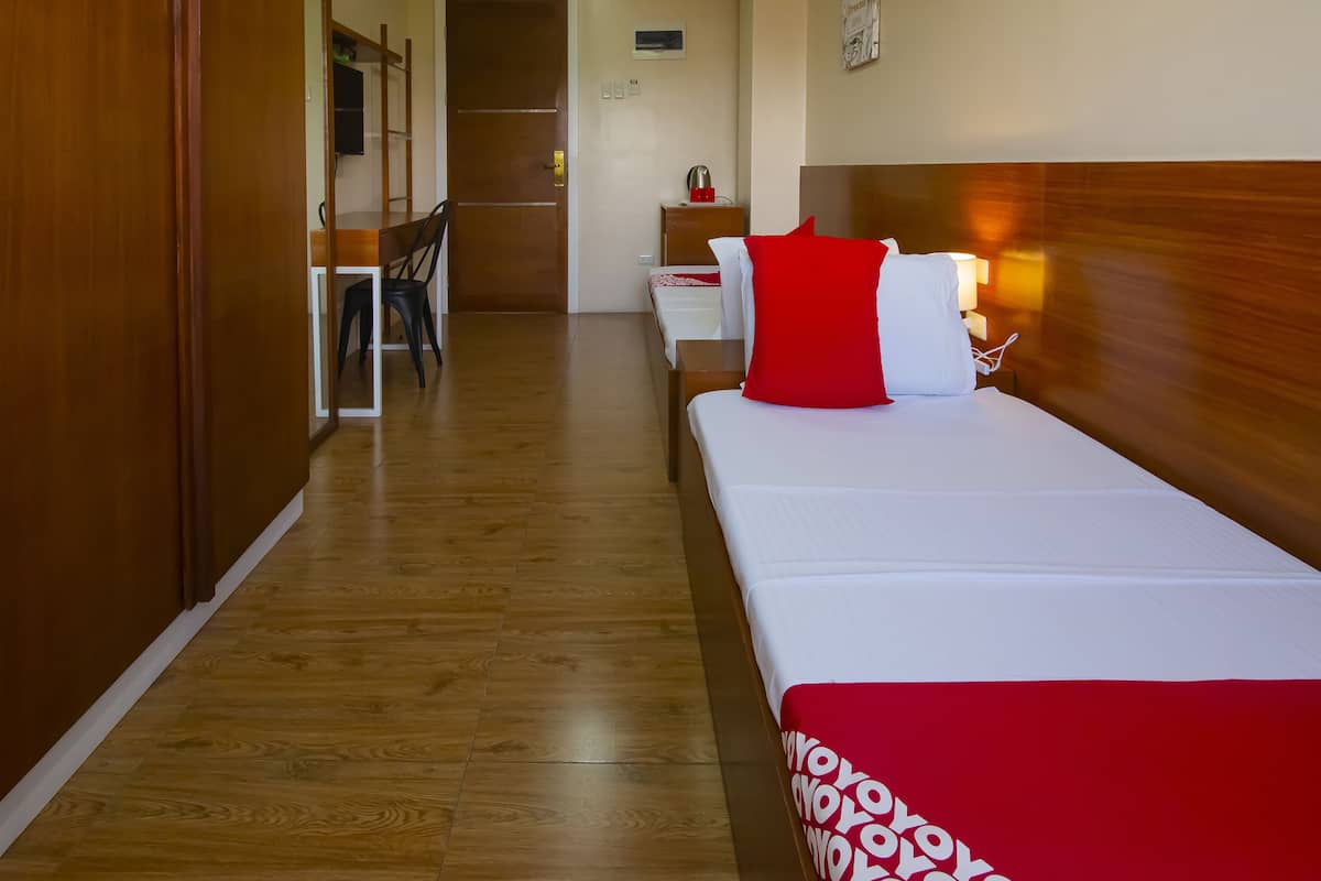 A hotel room with a wooden floor, a bed with a red and white pillow, a bedside table with a lamp, and a wooden wardrobe.