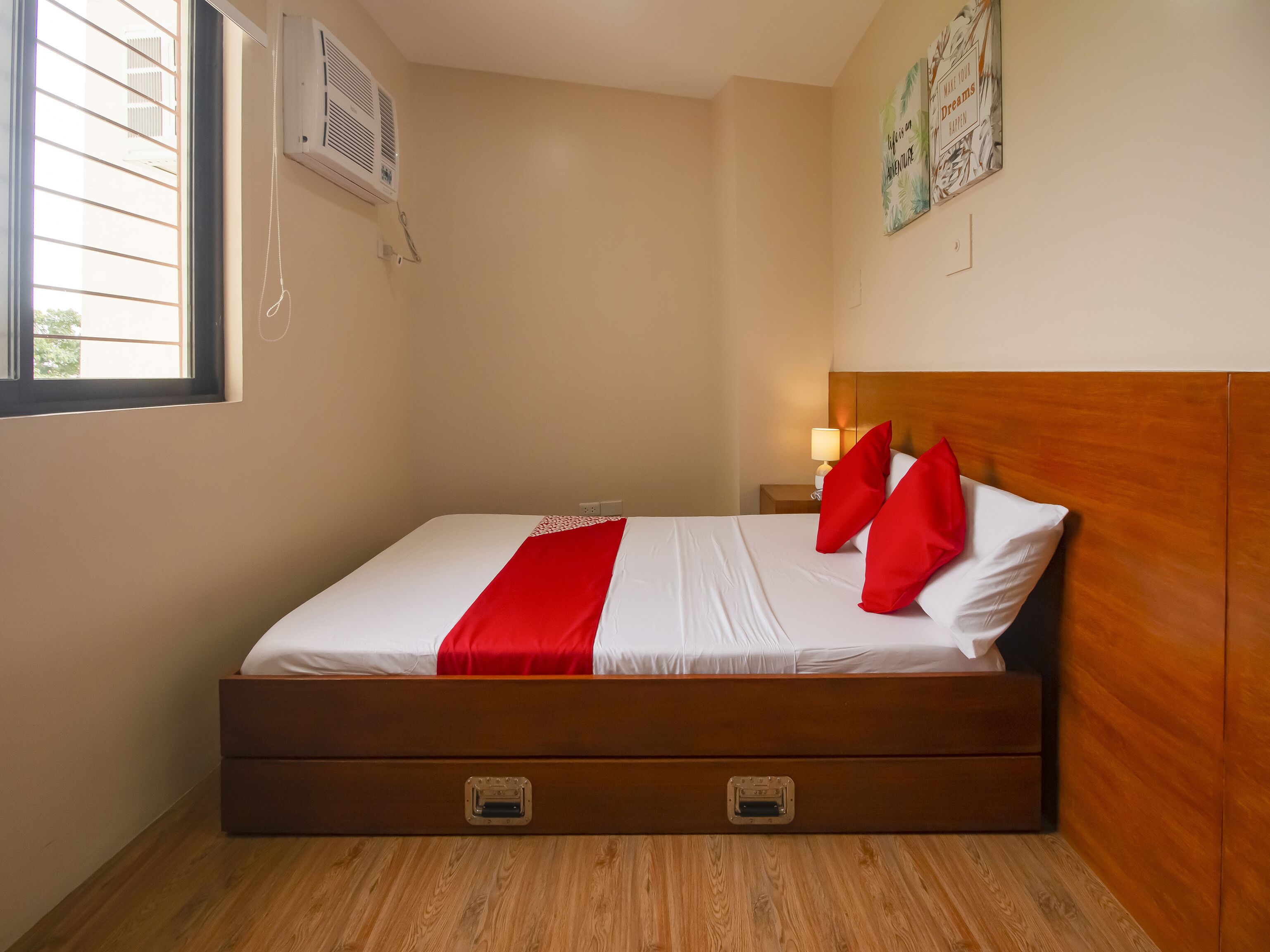 A neatly made bed with a wooden headboard, a red accent pillow, and a white comforter. There is a nightstand with a lamp and a wall-mounted air conditioner.