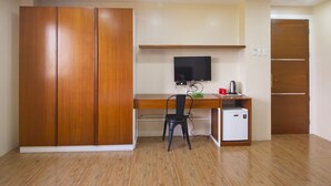 A room with a wooden wardrobe, a desk with a computer, a chair, a television, and a small refrigerator.