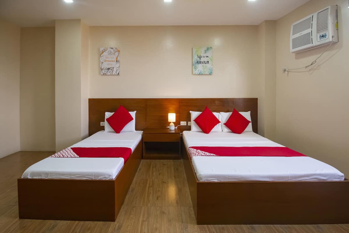 A hotel room with two beds, wooden headboards, red pillows, a bedside table with a lamp, and a wall-mounted air conditioner.