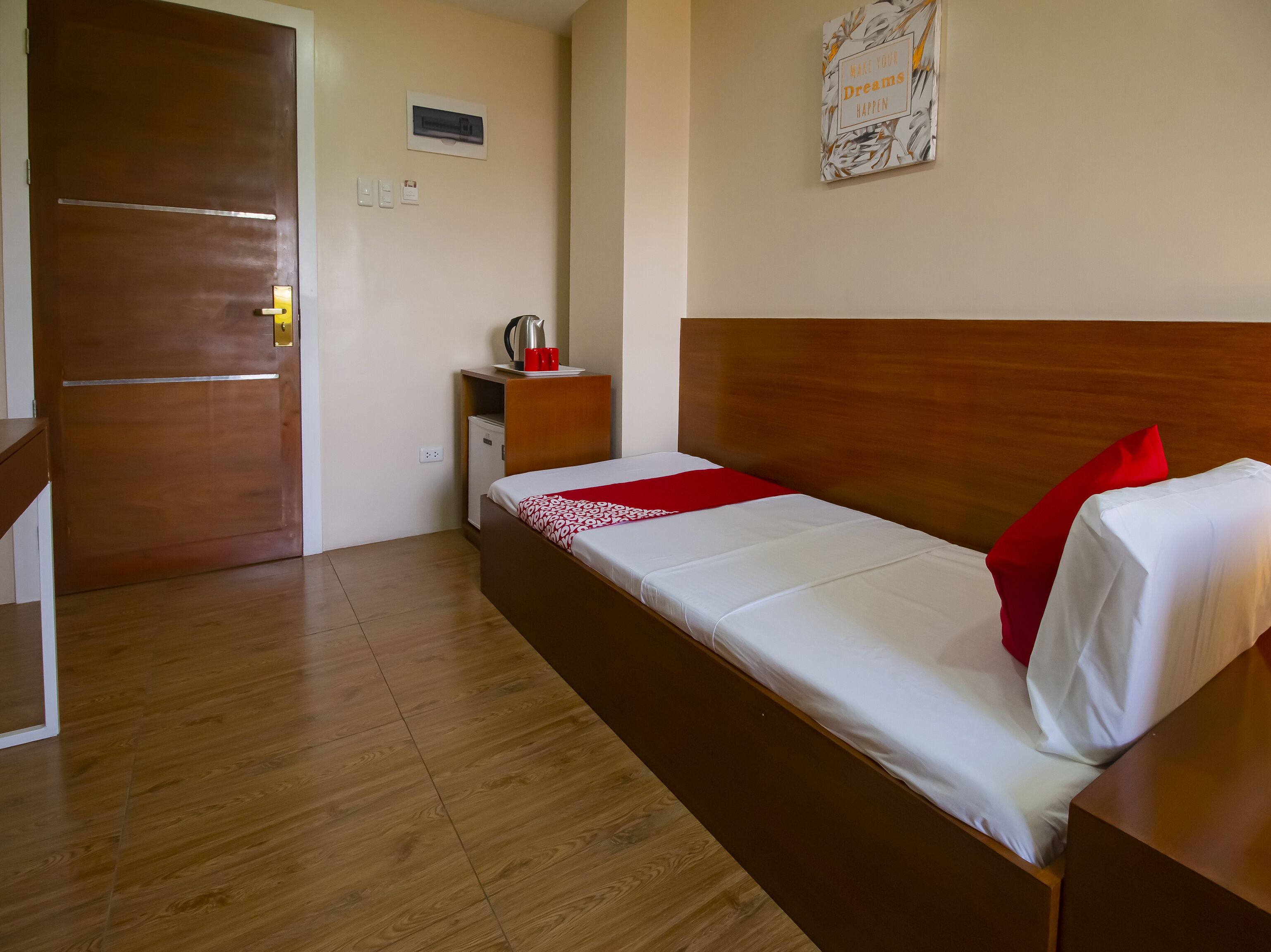 A hotel room with a wooden floor, a bed with white bedding and red pillows, a small bedside table with a kettle, a power outlet, and a wall-mounted air conditioner.