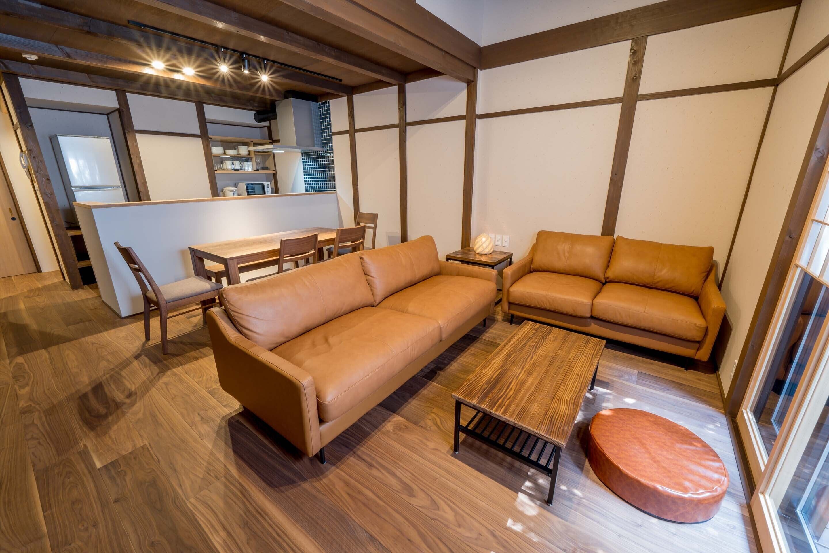 A modern living room with a wooden floor, a brown leather sofa, a wooden coffee table, and a small ottoman.