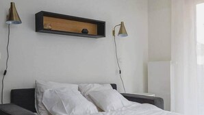 Una cama bien hecha con sábanas blancas y un cabecero oscuro en una habitación con pared blanca. Hay dos lámparas fijadas a la pared a cada lado de un estante de madera.