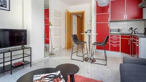 Una sala de estar moderna con televisor, asientos y una cocina con armarios rojos.