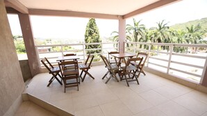 Un espacio de estar techado al aire libre con una mesa y sillas, vista a palmeras y colinas, y piso de baldosas.