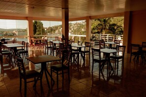An indoor dining area with tables and chairs, large windows, and a view of a body of water and buildings. - Pousada Alpes de Triunfo (Triunfo)