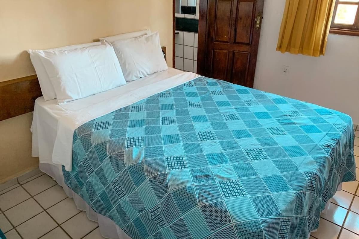 Uma cama com colcha azul estampada e almofadas brancas, cabeceira de madeira e piso de azulejos.