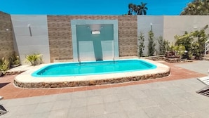 Una piscina con borde de piedra, rodeada por una zona de patio con plantas en macetas y un edificio moderno al fondo.