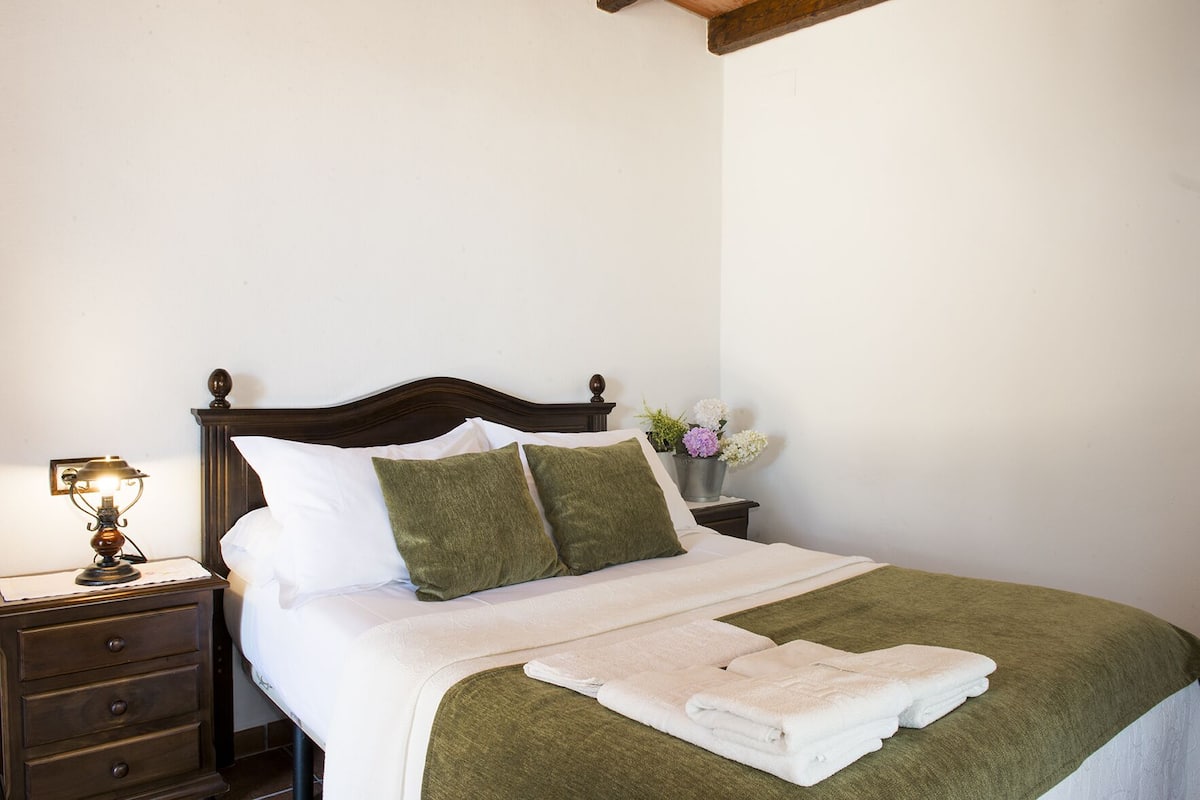 Uma cama bem arrumada com edredom branco, duas almofadas verdes e uma toalha branca dobrada.