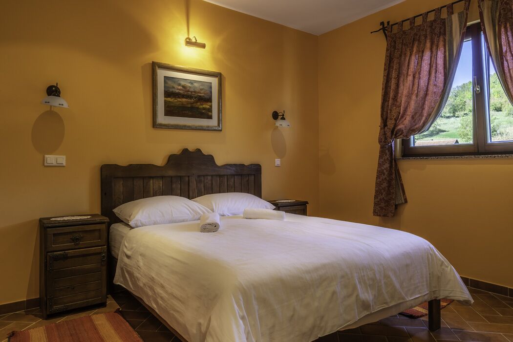 A bedroom with a wooden bed, white bedding, a painting on the wall, and a window with curtains.