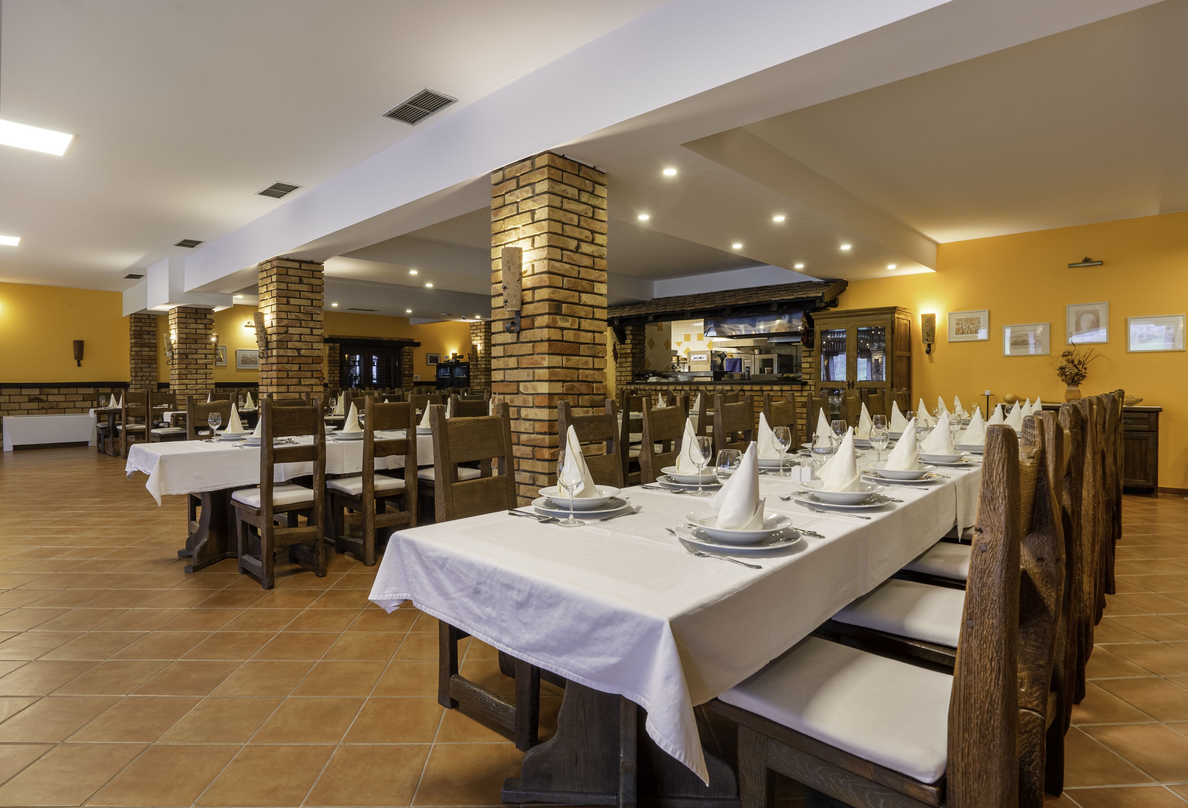 Um restaurante com mesas compridas dispostas para refeições, pilares de tijolo e uma área de cozinha ao fundo.
