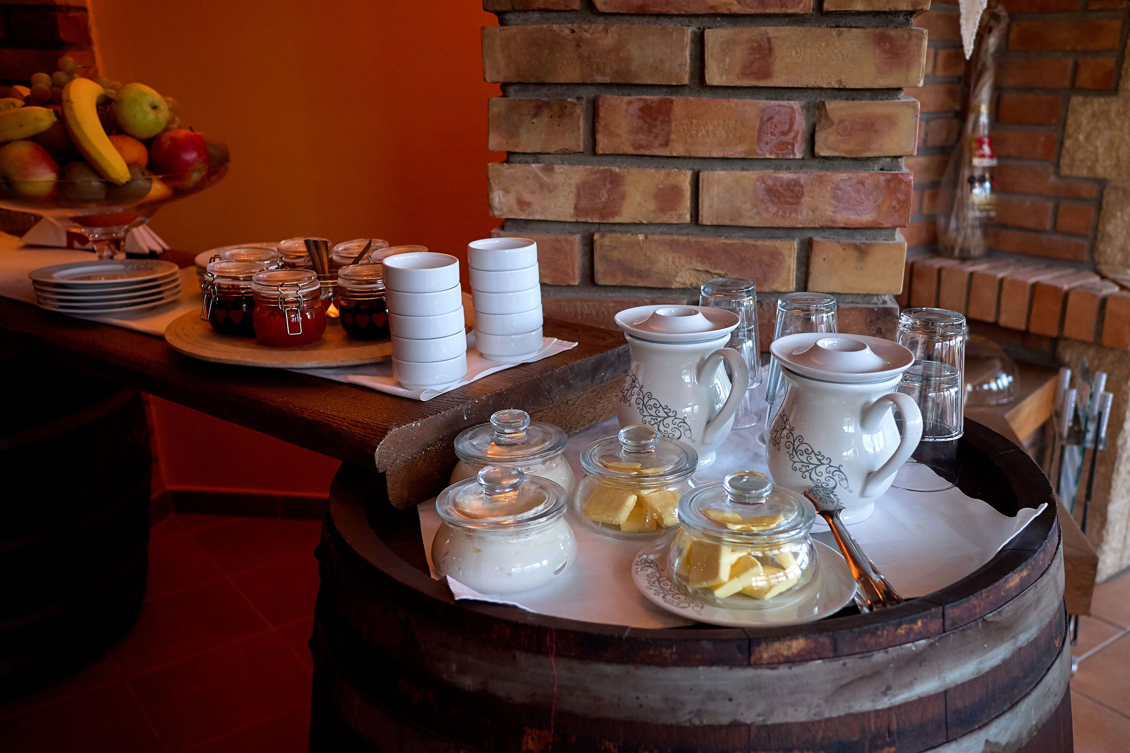 A breakfast buffet with a variety of food items and beverages, including fruits, jams, and dairy products, arranged on a wooden table with a brick wall background.
