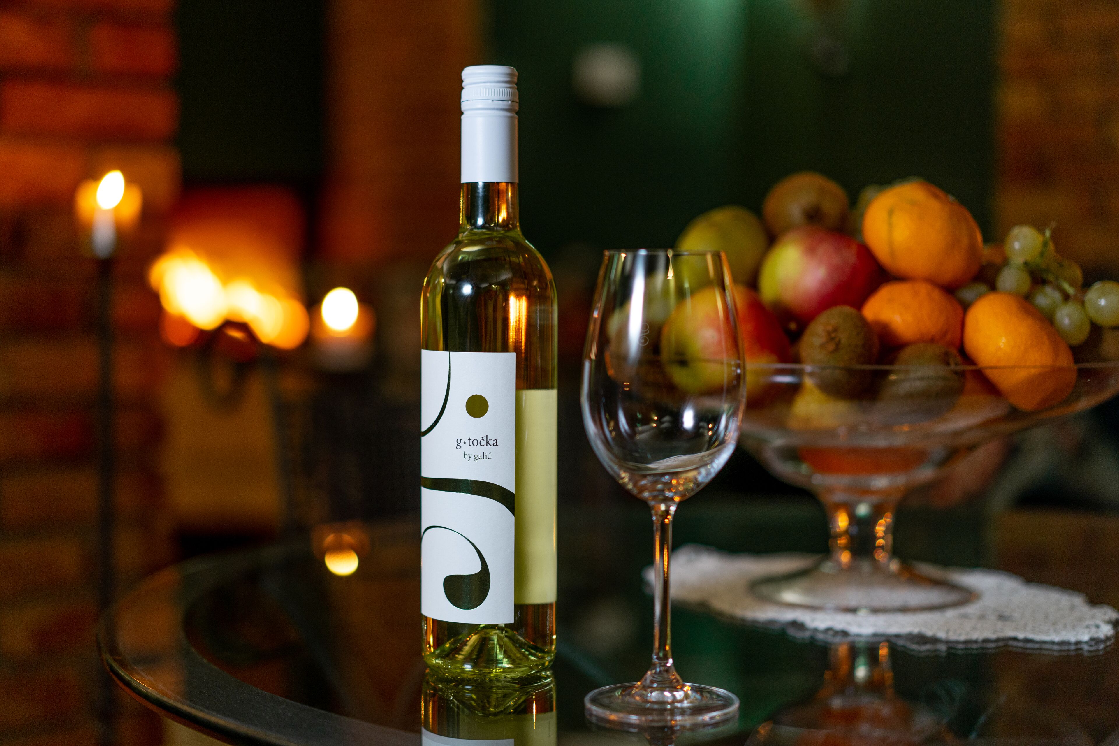 A bottle of wine, a wine glass, and a fruit bowl on a table with a glass top.