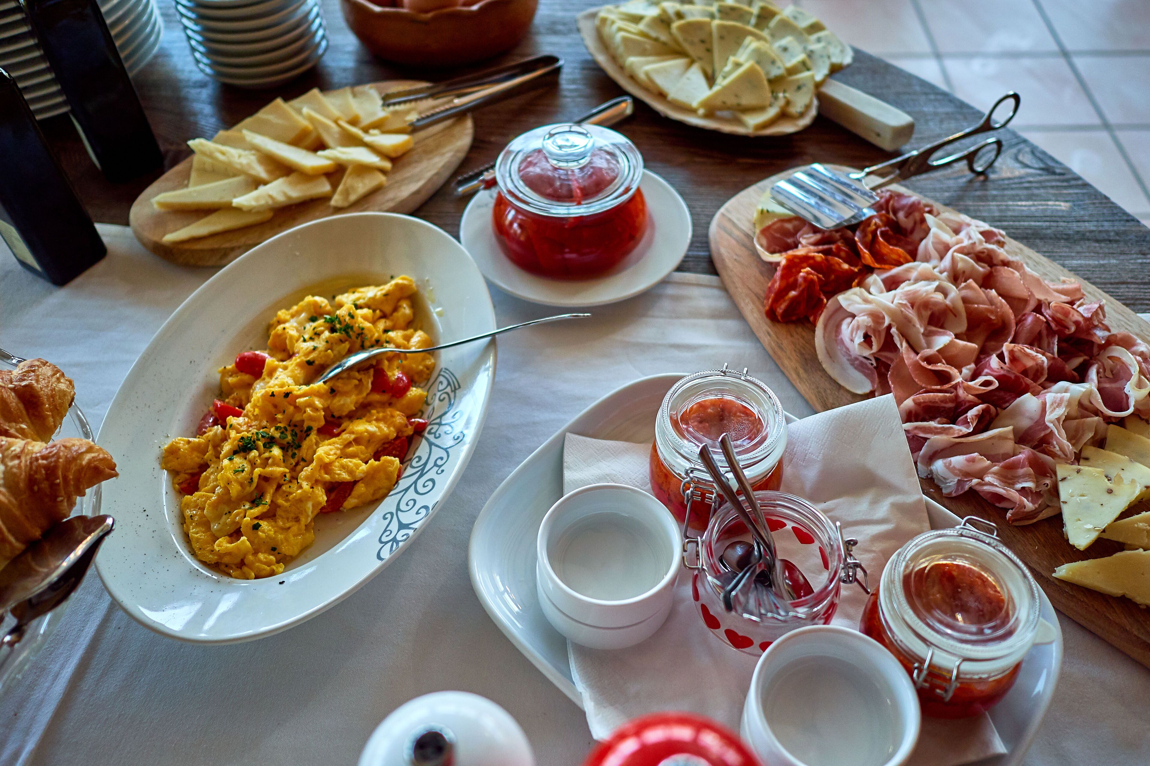 Un desayuno con huevos revueltos, queso y embutidos sobre una mesa de madera.