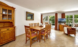 A dining area with a wooden table and chairs, a wooden cabinet with glass doors, a painting on the wall, and a large window with curtains. - Landhaus Pitzner (Garmisch-Partenkirchen)