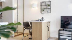 Un espacio habitable compacto con kitchenette, un comedor con una silla, un televisor sobre un mueble bajo y una estantería con plantas.