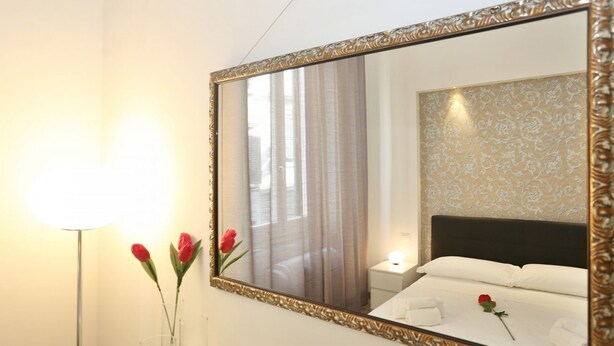 Ein Hotelzimmer mit großem Spiegel, einer Nachttischlampe, einer Vase mit Blumen und einem Bett mit weißer Bettwäsche.