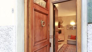 Una puerta de madera con manija metálica, que da paso a un cuarto con alfombra estampada, sofá y estante fijado a la pared con objetos decorativos.