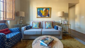 Un salón con un sofá, un sillón estampado, una mesa de centro con libros, un cuadro en la pared y dos lámparas sobre mesitas de noche.