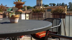 Una terraza en la azotea con una mesa redonda de metal y sillas, una maceta de terracota grande con plantas, y vistas a edificios y árboles.