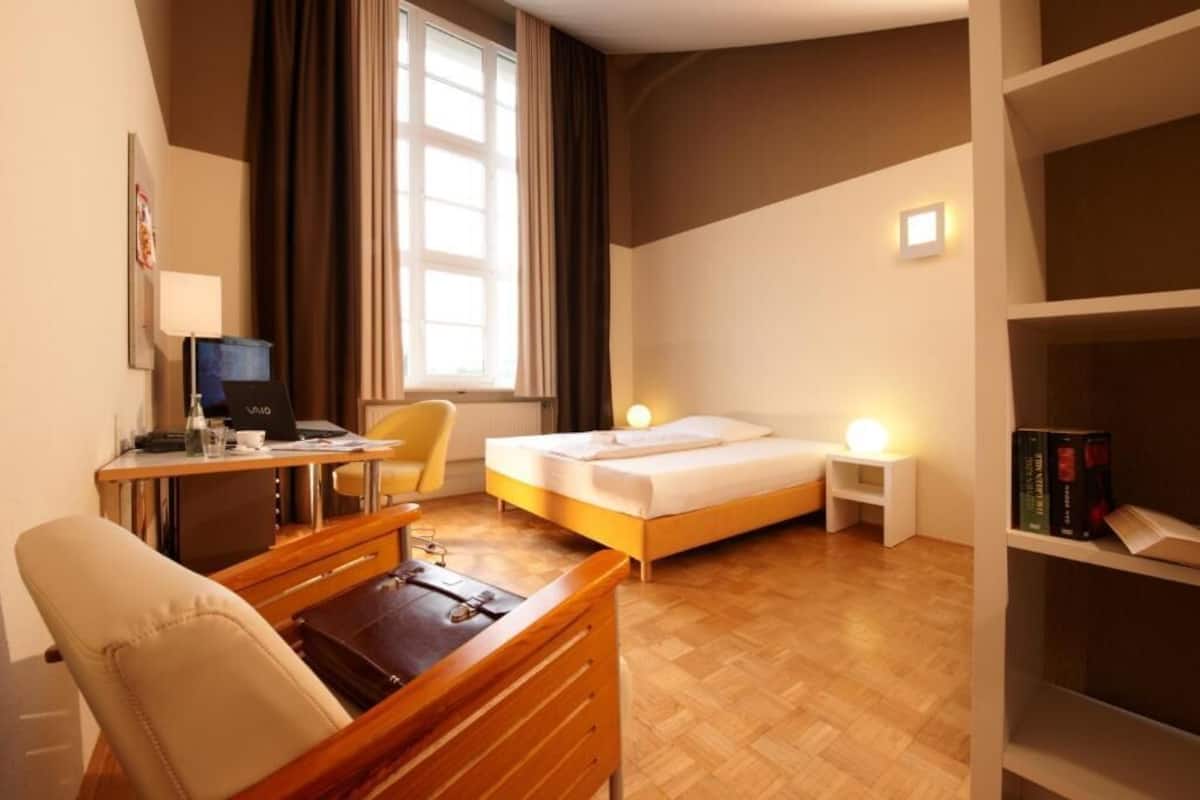 Ein Hotelzimmer mit einem Bett, einem Schreibtisch mit Computer, einem Sessel, einem Nachttisch mit Lampe und einem Bücherregal.