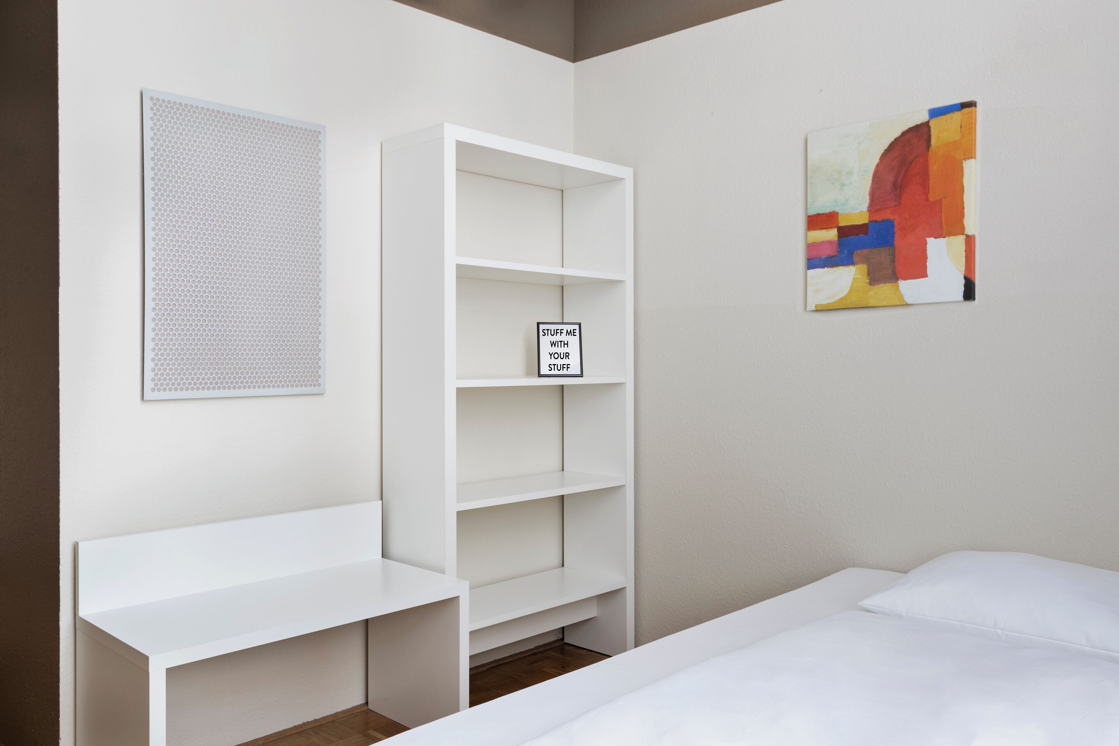 A room with a bed, a white shelving unit, a wall-mounted artwork, and a window with blinds.