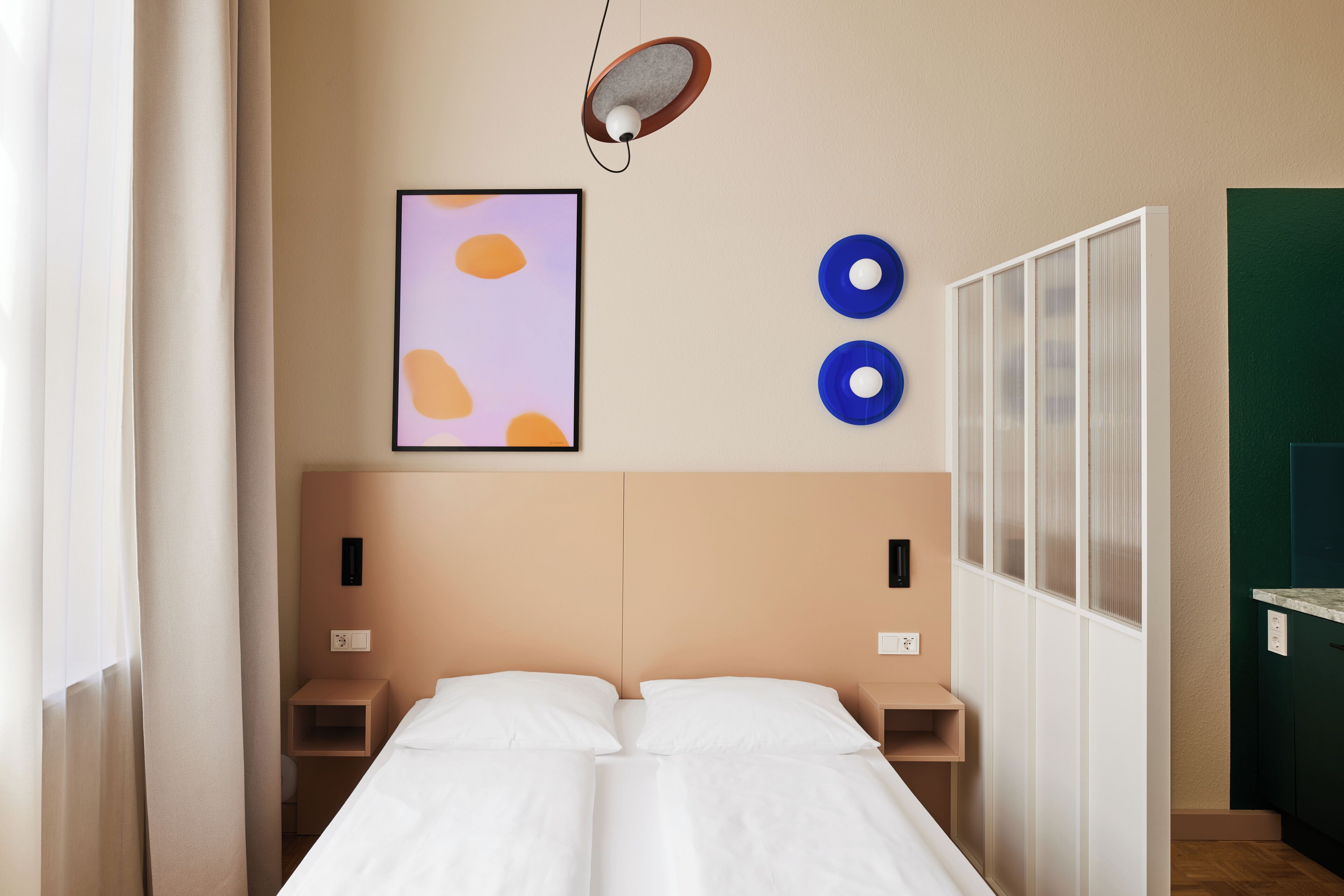 Ein Hotelzimmer mit Bett, Nachttischen, einer Wandleuchte, einem gerahmten Kunstwerk und einem Fenster mit Vorhängen.