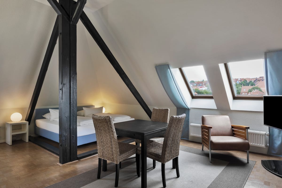 Ein modernes Schlafzimmer im Loft-Stil mit einem Bett, einem Esstisch und Stühlen. Ein Fernseher und ein Fenster mit Blick auf die Stadt sind vorhanden.