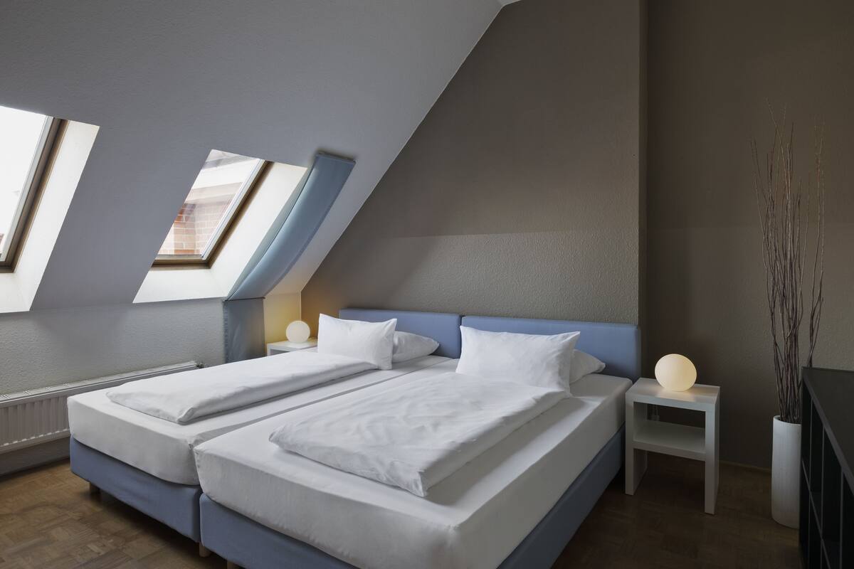 Ein modernes Schlafzimmer mit zwei Betten, Oberlichtern, einem Nachttisch mit Lampe und einer Vase mit Zweigen.