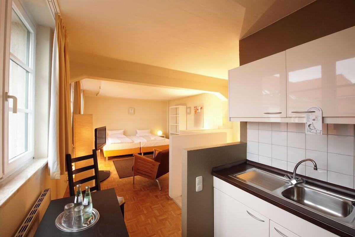 Ein kleines Hotelzimmer mit Küchenzeile, einem Bett, einer Sitzecke und einem Fenster mit Vorhängen.