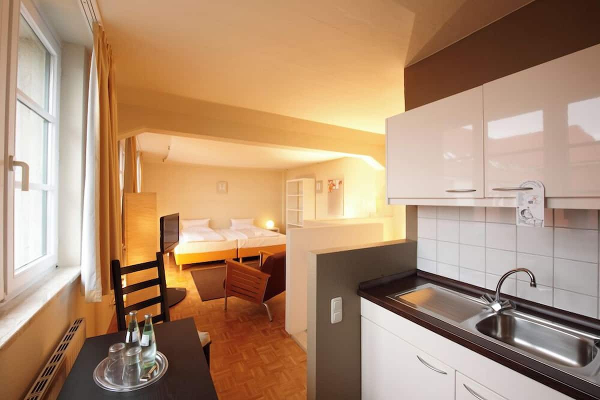 Habitación de hotel compacta con kitchenette, cama, pequeño rincón de descanso y ventana con cortinas.