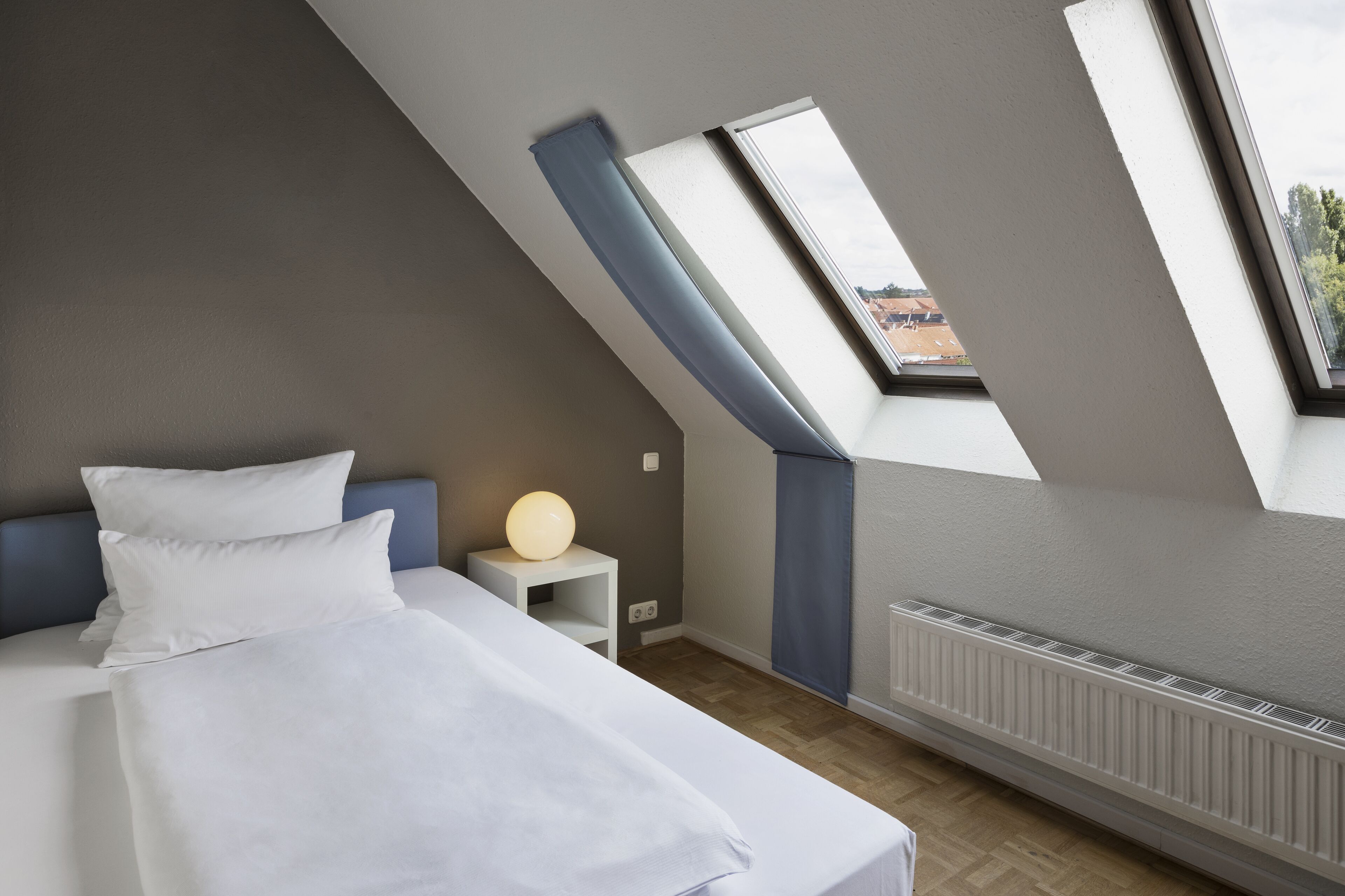 A modern bedroom with a sloped ceiling, a skylight, a bed with white linens, a bedside table with a lamp, and a radiator.